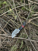 The Texan Knives Premium Custom Handmade Damascus Steel Guthook Skinning Knife with a resin handle and 7.5” blade rests open on dry twigs and green leaves, accompanied by its leather sheath.
