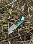The Texan Knives Elite Custom Handmade Damascus Steel Guthook Skinning Knife features a 7.5” patterned blade and a blue-green resin handle with star designs and a brass emblem, resting on dry branches outdoors. Includes premium leather sheath.