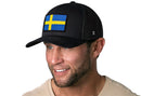 A man with a trimmed beard and mustache smiles, wearing the Haka Hat Swedish Flag Trucker Hat | Black Sweden Snapback by Haka Hat and a light shirt, posed against a white background.