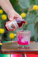 A hand pours vibrant magenta syrup from a Bottle of Brushfire Farms Prickly Pear Simple Syrup into a glass filled with ice and finished with a spiced rim. The glass rests on a wooden board, with vivid fabric and yellow flowers softly blurred in the background.