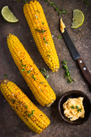 Three grilled corn on the cob garnished with herbs and SALTOPIA Infused Sea Salts from the Saltopia Mexican Food Lovers Set - TACO'CLOCK, served with seasoned butter, a knife, and lime wedges on a rustic surface.