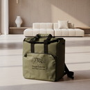 A Taiga Olive Back Pack Cooler by Taiga Coolers with black straps sits on the floor of a modern living room, with a white sofa and decorative vase in the background.