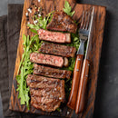 Sliced medium-rare steak served on arugula atop a wooden board, seasoned with SALTOPIA Infused Sea Salts’ “Saltopia Best Grilling Salts: Thrills for the Grill” and black pepper, with a fork and steak knife beside it.