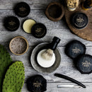 Beard care products from The Immaculate Beard, including a Shave Soap Puck for sensitive skin, a shaving brush, a straight razor, a cactus, a bowl, and various tins are arranged on a rustic wooden surface.