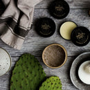 Flat lay of The Immaculate Beard Immaculate Natural Beard Balm For Men With Lightweight Hold tins, alongside a cup of coffee, a green cactus pad, a shaving brush, a gray towel, and a white bowl on a wooden surface. The rustic, masculine arrangement highlights the essentials of men’s grooming and the nourishing qualities of premium beard conditioning.