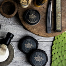 Flat lay of beard grooming products, featuring beard balm tins, The Immaculate Beard Shave Soap Puck—a natural shave soap ideal for sensitive skin—alongside a shaving brush, straight razor, and a box labeled Bismarck Razor, all arranged on a rustic wooden surface with a piece of cactus.