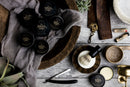 A rustic flatlay featuring The Immaculate Beard’s Conditioning Natural Beard Balm, shaving tools, a shaving brush, a cup of coffee, succulents, and brass dishes arranged on a wooden surface with gray cloth.