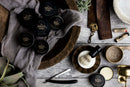 A flat lay of men’s grooming essentials, featuring The Immaculate Beard’s Immaculate Natural Beard Balm For Men With Lightweight Hold for superior beard conditioning, a shaving brush, a straight razor, a mug of coffee, plants, and grooming tools on a rustic wooden surface. Products are neatly arranged with earthy tones.
