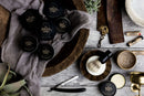 Grooming products, including a shaving brush, straight razor, and The Immaculate Beard’s Immaculate Dense Shave Soap For Smooth Men Wet Shaving for sensitive skin, plus nourishing beard balms, are arranged on a wooden table with cloth, plants, a cup of coffee, and small tins of The Immaculate Beard products—perfect for those who appreciate a traditional wet shave.