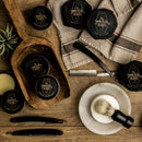 Flat lay of The Immaculate Beard grooming products featuring the Shave Soap Puck, along with beard balms, elegantly arranged on wood beside a shaving brush, bowl, straight razors, and linen cloth—ideal for anyone with sensitive skin.