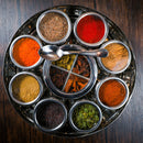 The Spice Wheel Indian Spice Wheel by Spice Wheel features a round container with nine compartments, each filled with vibrant spices. A small metal spoon is included and rests atop the spice wheel, which sits elegantly on a dark wooden surface—perfect for anyone looking to create restaurant-quality Indian cuisine at home.