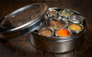 The Spice Wheel Indian Spice Wheel by Spice Wheel is a round stainless steel spice box with a glass lid, featuring multiple small containers for vibrant spices and a handy spoon. Placed on a wooden surface, this airtight spice container is perfect for keeping your ingredients fresh and preparing restaurant-quality Indian food at home.