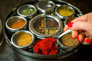 A hand with red nail polish holds a spoonful of red spice over the Spice Wheel Indian Spice Wheel by Spice Wheel—a round metal spice box featuring colorful spices in individual compartments, perfect for keeping your ingredients fresh in this airtight container and creating restaurant-quality Indian food at home.