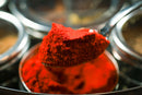 A close-up of a metal spoon holding a heap of vibrant red powdered spice, likely paprika or chili powder, above the airtight Spice Wheel Indian Spice Wheel container filled with more of this essential spice—perfect for preparing restaurant-quality Indian food.