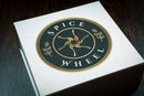 A white, airtight Spice Wheel Indian Spice Wheel container from the brand Spice Wheel rests on a dark wooden surface, showcasing its circular black and gold logo with abstract spice graphics and decorative wheat accents—an ideal Indian spice box for anyone looking to bring restaurant-quality Indian food into their kitchen.