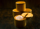 The Spice Wheel Bamboo Salt Container filled with Pink Himalayan Salt, complete with a wooden spoon, stands open and surrounded by three closed Spice Wheel containers on a dark wooden surface.