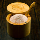 The Spice Wheel Bamboo Salt Container, filled with Pink Himalayan Salt and featuring a matching wooden lid and small wooden spoon, rests elegantly on a dark wooden surface.