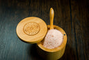The Spice Wheel Bamboo Salt Container, filled with Pink Himalayan Salt and paired with a small wooden spoon, rests elegantly on a dark wooden surface. The open lid reveals a beautifully engraved Spice Wheel logo and text.