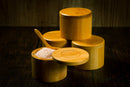 A stack of cylindrical bamboo spice containers sits on a dark wooden surface. One Spice Wheel Bamboo Salt Container, filled with Pink Himalayan Salt and accompanied by a small wooden spoon, is open with its lid resting nearby. Each container features the Spice Wheel logo branded on top.