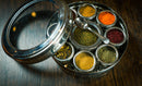 A Spice Wheel Mediterranean Spice Wheel by Spice Wheel, featuring a round stainless steel box with a transparent lid, holds small containers filled with an array of vibrant Mediterranean spices and herbs. A small metal spoon rests inside, and the set is displayed on a dark wooden surface.