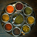 Overhead view of the Spice Wheel Mediterranean Spice Wheel by Spice Wheel—a circular stainless steel spice box featuring seven small containers, each filled with a variety of Mediterranean spices like powders, dried herbs, whole cloves, and green cardamom pods. A small metal spoon rests in the central compartment atop a wooden surface.
