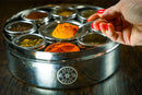 A hand with red nails holds a spoon of orange spice above the Spice Wheel Mediterranean Spice Wheel by Spice Wheel, a round stainless steel spice box filled with an array of colorful Mediterranean spices, beautifully displayed on a wooden surface.