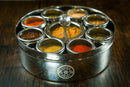 A round metal Spice Wheel Mexican Spice Wheel by Spice Wheel, featuring nine small containers filled with vibrant Mexican spices and a small spoon in the center, sits on a dark wooden surface.