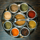 The Spice Wheel Mexican Spice Wheel by Spice Wheel is a round metal spice box, inspired by the traditional Mexican Spice Wheel. It features nine compartments filled with a variety of colorful Mexican spices, with a small metal spoon placed in the center compartment. The vibrant display sits atop a dark wooden surface.