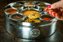 A hand with red nails holds a spoon of ground spice above the Spice Wheel Mexican Spice Wheel by Spice Wheel, featuring an array of colorful spices in small containers, all arranged on a wooden surface.