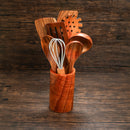 A wooden container holds assorted wooden kitchen utensils, including pieces from the Spice Wheel Acacia Wooden Spoon Set by Spice Wheel and a metal whisk, set against a dark wooden background.
