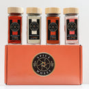 Four glass spice jars with wooden lids sit atop an orange Spice Wheel box. The Spice Wheel Foundations of Flavor Gift Set by Spice Wheel features four essential spices: Hungarian paprika, white pepper, smoked paprika, and Himalayan pink sea salt.