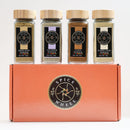 Four glass jars of aromatic spices—anise, clove, fennel, and lavender—from the Spice Wheel Baking Botanicals Gift Set are arranged on the bright orange Spice Wheel box featuring a circular black-and-gold logo, making it perfect for floral baking creations.