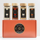 Four spice jars labeled Vanilla Sugar, Cinnamon Sugar, Ginger Sugar, and Caramel Sugar are arranged atop an orange Spice Wheel Cozy Sugar Gift Set box featuring the Spice Wheel’s circular logo—a delightful collection of flavored sugars perfect for gifting.