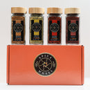 Four glass jars of Mediterranean spices—Brown Za’atar, Green Za’atar, Aleppo Pepper, and Sumac—are displayed atop the orange Spice Wheel Mediterranean Spice Essentials Gift Set box, featuring a circular logo. This beautifully arranged set by Spice Wheel is perfect for enhancing your cooking with authentic Mediterranean flavors like za’atar and more.