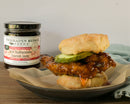 A golden fried chicken biscuit sandwich with pickles is served alongside a jar of Terrapin Ridge Farms Hot Habanero Bacon Jam—ideal as a savory charcuterie board topping or spread for saucy chicken sandwiches.