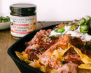 A skillet of nachos topped with pulled pork, jalapeños, and sauce sits beside a jar of Terrapin Ridge Farms Terrapin Ridge Hot Habanero Bacon Jam—a sweet and spicy charcuterie board topping. A small bowl of green garnish appears in the background.