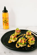 A black plate holds five sushi pieces topped with diced tuna, avocado, jalapeños, and a drizzle of Terrapin Ridge Farms Sriracha Aioli Garnishing Squeeze. A bottle of Terrapin Ridge Farms sauce and chopsticks are nearby.