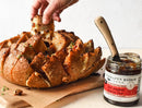 A hand pulls a cheesy slice from a loaf, revealing melted cheese and herbs. Beside it sits an open 2oz jar of Terrapin Ridge Farms Hot Pepper Bacon Jam with a knife inside.
