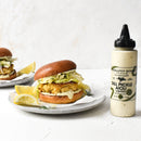 A chicken burger with lettuce and sauce sits on a plate with lemon wedges beside a bottle of Terrapin Ridge Farms Dill Pickle Aioli Squeeze—a zesty condiment by Terrapin Ridge Farms. Another burger is in the background, all on a white surface.