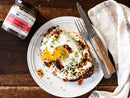 A plate with toast and a fried egg, garnished with herbs, sits beside a jar of Terrapin Ridge Farms Hot Pepper Bacon Jam—ideal for charcuterie boards—along with a fork, knife, and striped cloth napkin.