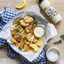 Crispy fried potato wedges topped with Terrapin Ridge Farms Dill Pickle Aioli Squeeze and fresh dill, served with lemon wedges. Nearby are seafood sauce, a bottle of aioli, a salt bowl, and a blue checked napkin.