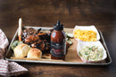 A tray with barbecue ribs, two dinner rolls, pickle slices, macaroni and cheese, coleslaw, and a bottle of Terrapin Ridge Farms Hot Pepper Bacon Grill & Wing Squeeze on a wooden table with a red plaid napkin.