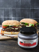 Two large sandwiches with lettuce, tomato, cheese, and sauce on ciabatta buns sit on a wooden board. In front is a jar of Terrapin Ridge Farms Hot Pepper Bacon Jam. A black tiled wall is in the background.