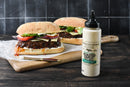 Two plant-based burgers with lettuce, tomato, onion, and cheese sit on a wooden board next to a bottle of Terrapin Ridge Farms Avo Aioli Roasted Garlic Squeeze. A knife with aioli rests nearby on parchment paper.