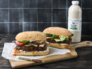 Two gourmet burgers with lettuce, tomato, cheese, onions, and sauce sit on a wooden board beside a bottle of Terrapin Ridge Farms Roasted Garlic Avo Aioli, against a dark tiled background. A knife with aioli rests nearby.