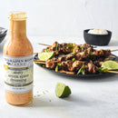 A bottle of Terrapin Ridge Farms Zesty Seven Spice Dressing sits on a white surface near grilled meat skewers, lime wedges, and a small bowl of rice, with a light background.
