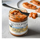 A jar of Terrapin Ridge Farms Bleu Cheese Stuffed Olive Dip sits on a table with a black spreader—a perfect Charcuterie Board Spread for toasted bread slices in the blurred background.