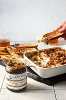 A hand dips bread into a cheesy baked dish topped with onions and bacon. An open jar of Terrapin Ridge Farms Balsamic Onion Jam, the perfect cheese board condiment, sits in front. Crackers and drinks are visible in the background.