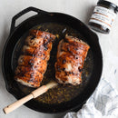 Two glazed pork tenderloins in a cast iron skillet with a basting brush, Terrapin Ridge Farms Blueberry Bourbon Pecan Jam - 2oz, and a white kitchen towel nearby.