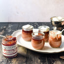 Four small glass jars of chocolate pudding, some with whipped cream, are arranged on a white platter. Next to them on a dark wooden table is a jar of Terrapin Ridge Farms Raspberry Amaretto Jam with a spoon.